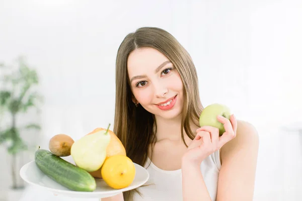 Joven mujer sonriente sosteniendo surtido de frutas servidas en plato blanco y manzana verde sobre fondo claro. Sonrisa sana, hermosa, concepto de belleza —  Fotos de Stock