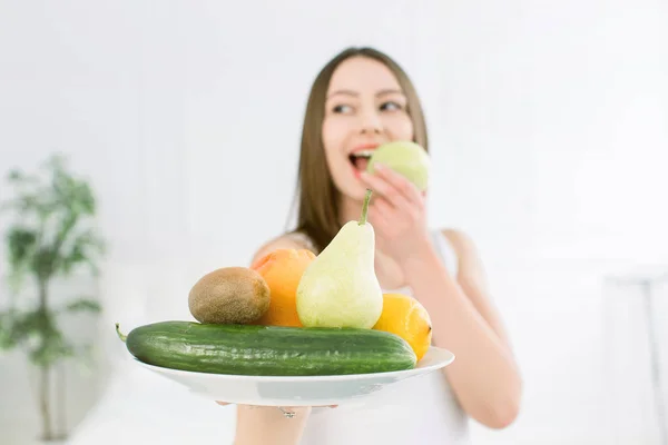 Mujer sosteniendo frutas surtido servido en plato blanco sobre fondo claro. Retrato de cerca de una joven alegre con dientes perfectos mordiendo manzana verde. Concepto de belleza saludable —  Fotos de Stock