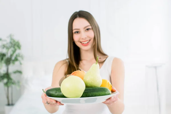 Hermosa joven sonriente sosteniendo un plato de frutas frescas, manzana, pera, limón, pomelo, kiwi y pepino. Alimentación saludable, concepto de belleza — Foto de Stock