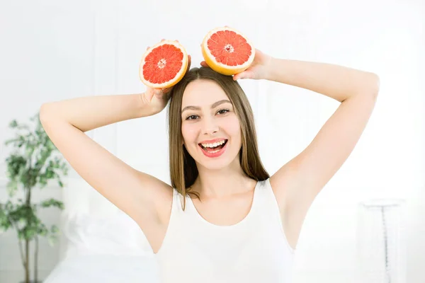 Retrato de una alegre y atractiva chica bastante positiva con dos piezas de pomelo en las manos sobre la cabeza sobre el fondo claro. Concepto de comida saludable. Cuidado de la piel y belleza. Vitaminas y minerales . — Foto de Stock