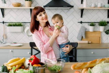 masada evde mutfak ayakta tutan küçük bebek kız yemek taze yeşil elma, mutlu gülümseyen anne. Annelik, Aile, gıda, sağlıklı beslenme, yemek ve insanlar kavramı