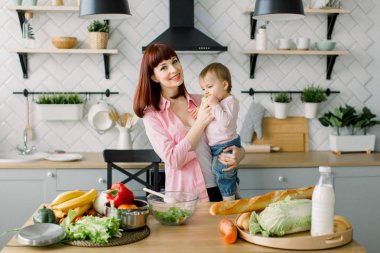 masada evde mutfak ayakta tutan küçük bebek kız yemek taze yeşil elma, mutlu gülümseyen anne. Annelik, Aile, gıda, sağlıklı beslenme, yemek ve insanlar kavramı