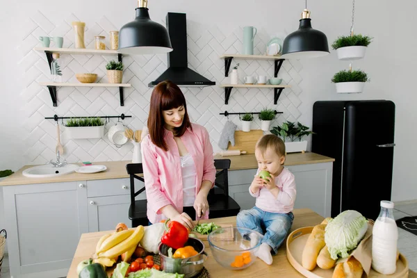 Fiatal boldog anya, darabolás, piros paprika és a baba kislány zöld alma a kezében ül otthon a Vacsoraasztal családi konyha, élelmiszer, egészséges táplálkozás, főzés, és anyák napja koncepció — Stock Fotó