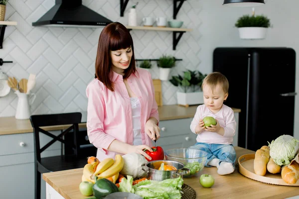 Young happy mother chopping red pepper and little baby girl holding green apples in her hands sitting on dinner table at home kitchen family, food, healthy eating, cooking and Mothers Day concept