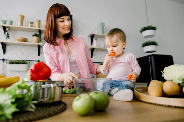 Mosolygó fiatal anya készül friss salátával, üvegtál állva a modern konyha asztalnál közeli portréja. Baby kislány enni a répát. Egészséges élelmiszer koncepció — Stock Fotó