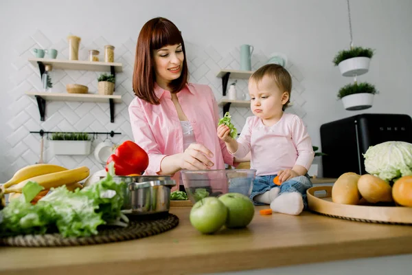 Mothers Day, food, healthy eating, cooking and family concept - happy mother and little baby girl preparing salad with fresh lettuce at home kitchen