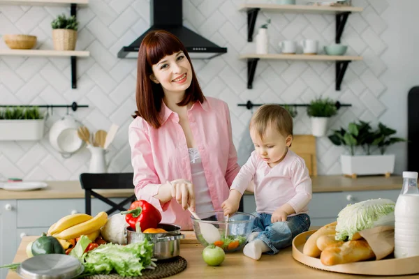Egy fiatal és gyönyörű anya rózsaszín ingben készül egy friss növényi saláta otthon a konyhában, ül az asztal kis aranyos lányával. anya és lánya — Stock Fotó