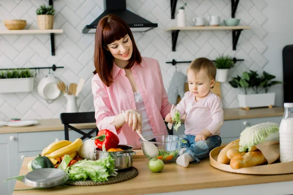 Egy fiatal és gyönyörű anya rózsaszín ingben készül egy friss növényi saláta otthon a konyhában, ül az asztal kis aranyos lányával. anya és lánya — Stock Fotó