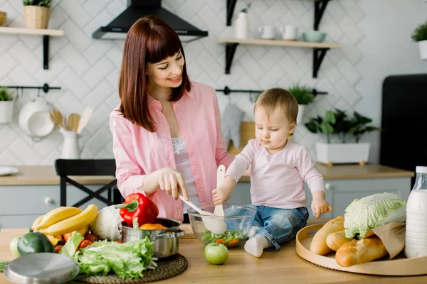 Bonito pequena menina bebê segurando garfo de madeira ajudando sua mãe bonita para misturar salada na mesa na cozinha. Jovem mulher bonita com filha cozinhar salada fresca em casa . — Fotografia de Stock