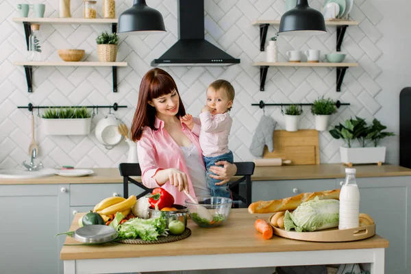 Cheerful beautiful mother in a pink shirt is preparing a fresh vegetable salad at home in the kitchen, holding her little cute daughter. Mother and daughter, healthy food concept