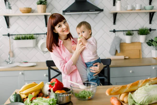 Feliz sorrindo mãe segurando menina bebê comendo maçã verde fresca, de pé na mesa em casa cozinha. Conceito de maternidade, família, alimentação, alimentação saudável, culinária e pessoas — Fotografia de Stock