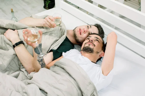Two handsome guys drink white wine. lying in bed at home. Young gay couple lying under blanket on bed in light room — Stock Photo, Image