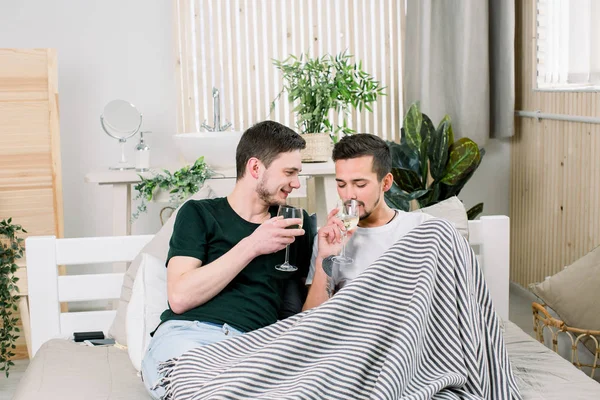 Счастливая пара геев лежит в постели с бокалами вина дома. Геи обнимаются и целуются в постели. Дом любви гей-пар — стоковое фото