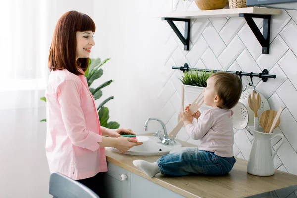 Genç mutlu anne ve sevimli kıvırcık yürümeye başlayan çocuk kızı beyaz bir mutfakta beraber eğleniyor. Kız bebek lavabonun oturan ve ahşap çatal ile oynarken annesi yemekleri, levha, yıkama — Stok fotoğraf