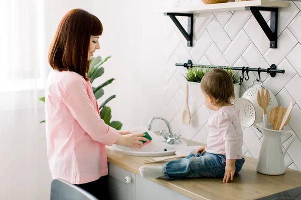 Genç mutlu anne ve sevimli kıvırcık yürümeye başlayan çocuk kızı beyaz bir mutfakta beraber eğleniyor. Kız bebek lavabonun oturan ve ahşap çatal ile oynarken annesi yemekleri, levha, yıkama — Stok fotoğraf