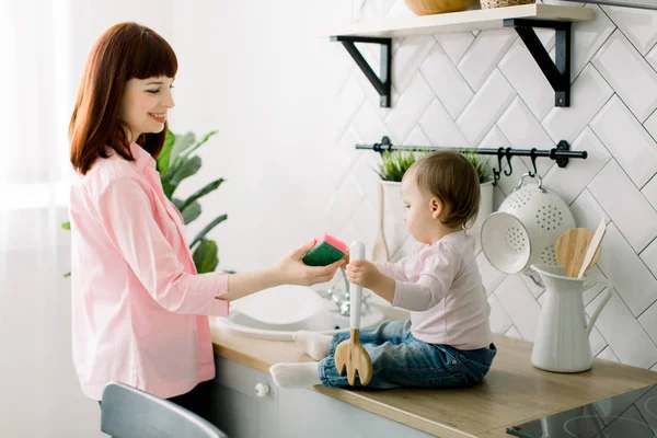 Babymeisje helpen haar mooie moeder afwas in de keuken. Focus op meisje. Een klein kind met belangstelling helpt haar moeder met huishoudelijke taken. Het meisje is blij om te leren om de afwas. — Stockfoto