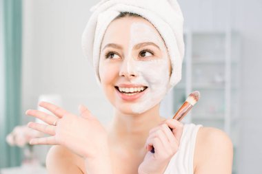 Mutlu ve duygusal Spa kız yüz maskesi uygulayarak. Estetik prosedür. Kız bir fırça ile yüzün yarısında kil bir anti-aging maskesi uygular