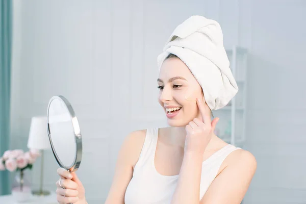 Retrato de atraente mulher branca sorridente no estúdio branco tiro limpar o rosto olhando para o espelho aplicando Foundation On Facial Skin — Fotografia de Stock