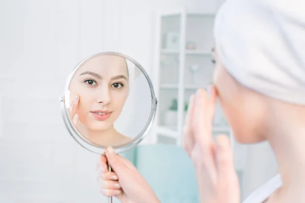 Όμορφη γυναίκα στο λευκό μπουρνούζι και πετσέτα που βλέπουν το τέλειο δέρμα της στον καθρέφτη και εφαρμόζοντας τονικό κρέμα βάσης στο πρόσωπο — Φωτογραφία Αρχείου