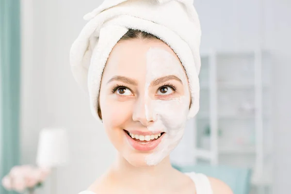 Een mooi jong meisje met een witte handdoek op haar hoofd een nuttige klei masker toegepast op de helft van het gezicht — Stockfoto