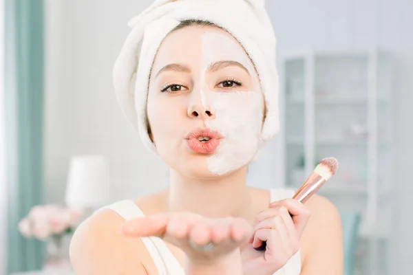 Young happy and smiling beautiful woman applying clay facial mask to half the face and sending an air kiss — Stock Photo, Image