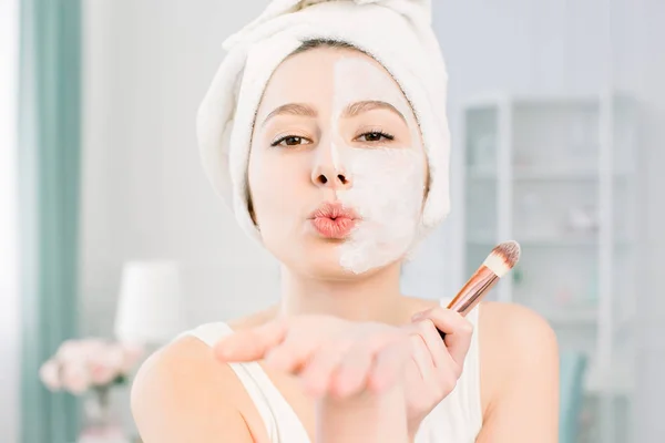 Jovem mulher bonita feliz e sorridente aplicando máscara facial de barro para metade do rosto e enviando um beijo de ar — Fotografia de Stock