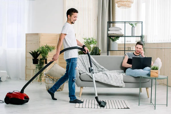 Couple of gay cleaning living room at home, Young gay man holding the laptop, sitting on sofa. Young gay using vacuum cleaner and cleaning the floor — Stock Photo, Image