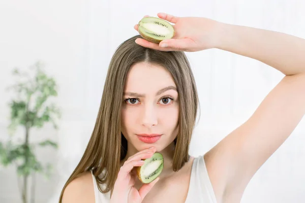 Mujer hermosa frunciendo el ceño mirando a la cámara con una ceja levantada siendo reflexiva. Joven chica bonita con dos rebanadas de kiwi en el fondo claro — Foto de Stock