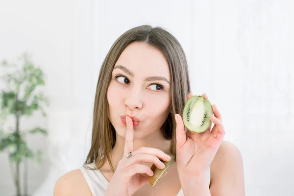 Retrato de belleza de una mujer muy sonriente con una piel sana mostrando un gesto de silencio con el dedo sobre los labios y sosteniendo rebanadas de kiwi, aisladas sobre un fondo claro — Foto de Stock