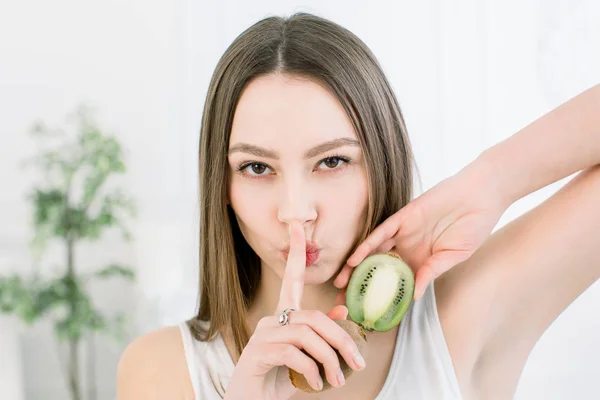 Retrato de belleza de una mujer muy sonriente con una piel sana mostrando un gesto de silencio con el dedo sobre los labios y sosteniendo rebanadas de kiwi, aisladas sobre un fondo claro — Foto de Stock