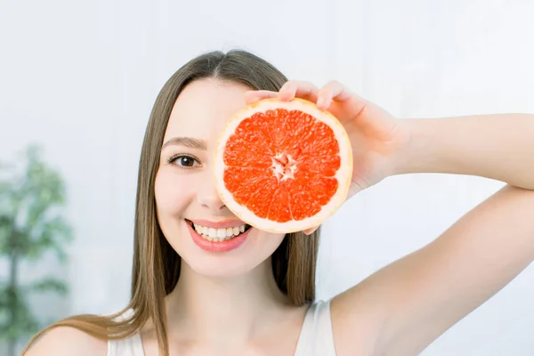 Primer plano de hermosa chica con el pelo castaño claro y pecas sonriendo labio mordedor, sosteniendo rebanada de pomelo, sobre fondo claro. Alimentación saludable, cuidado de la piel, concepto de belleza —  Fotos de Stock