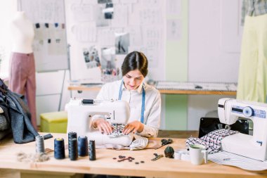 Onun çalışma yerinde otururken stüdyoda dikiş makinesi ile genç kadın dikiş. Moda Tasarımcısı dikkatle yeni moda stilleri oluşturma. Terzici giysi yapar