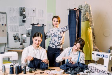 Genç gülümseyen moda tasarımcısı dikiş atölyesinde askılar üzerinde el yapımı pantolon ile poz ve kamera bakıyor. İki güzel terzi atölyesinde dikiş. Terzici çalışma