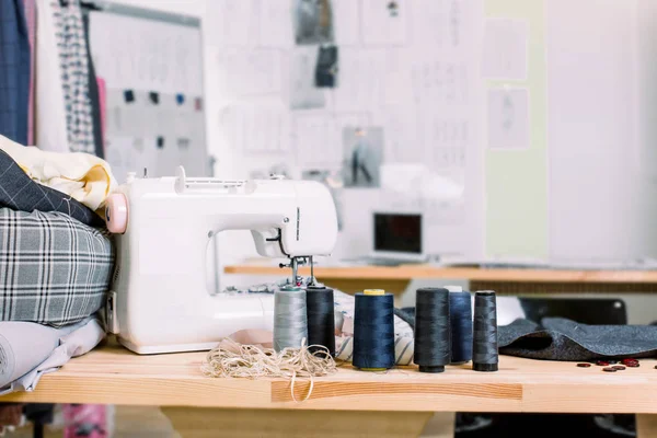 Μια φωτογραφία από ένα ηλιόλουστο στούντιο σχεδιασμού μόδας. Βλέπουμε ραπτομηχανή και διάφορα πράγματα που σχετίζονται με την ραπτική στο τραπέζι, πολύχρωμα υφάσματα, κρεμασμένα ρούχα. Χώρος εργασίας ράφτη με μοντέρνα ραπτομηχανή — Φωτογραφία Αρχείου
