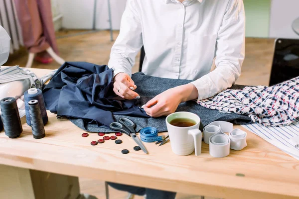 お針子の職場。ドレスメーカー手のトリミング画像ボタンを縫います。テーブルの上のボタン、ズボン、パターン、はさみ、糸および針、テープ測定および茶のための材料. — ストック写真