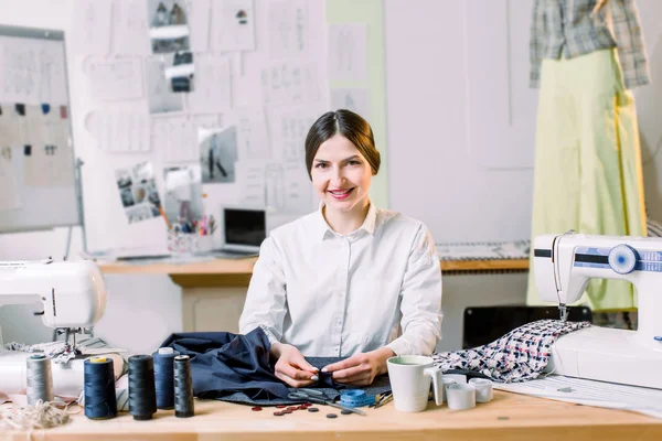 Χαμογελαστός θηλυκός σχεδιαστής μόδας κάθεται στο γραφείο του γραφείου. Μοδίστρα, ράφτης, έργα και ράφτρα concept-πορτρέτο του χαμογελαστή μόδας σχεδίαση χρησιμοποιώντας ραπτομηχανή — Φωτογραφία Αρχείου