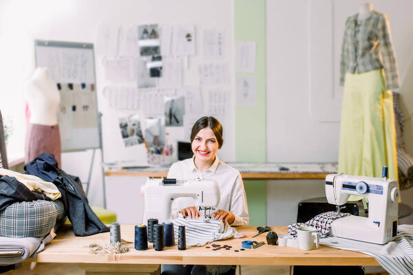 Beautiful seamstress sewing at her workshop. Dressmaker working on the sewing machine. Tailor making a garment in her workplace.