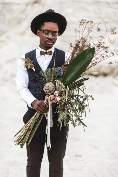 Pemuda kulit hitam Afro Amerika berdiri di belakang ngarai pasir yang indah saat matahari terbenam dan memegang buket pernikahan boho bergaya pedesaan. Pernikahan gurun, ngarai — Stok Foto