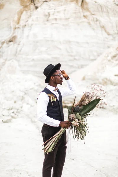 Knappe Afrikaanse man bruidegom in zwart pak en hoed Holding bruiloft boeket in rustieke stijl en staande in Desert Canyon tegen mooi landschap. — Stockfoto