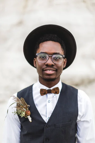 Close-up portret van knappe glimlachende Afro Amerikaanse bruidegom in zwarte hoed, wit overhemd en zwart zwart vest poseren in Canyon tegen prachtige woestijnlandschap bij zonsondergang. — Stockfoto