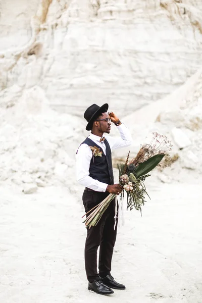 Knappe Afrikaanse man bruidegom in zwart pak en hoed Holding bruiloft boeket in rustieke stijl en staande in Desert Canyon tegen mooi landschap. — Stockfoto