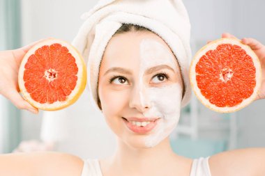 Hafif arka planda beyaz yüz maskesi ve greyfurt halfs ile güzel genç kadın. Elinde bir greyfurt tutan kafasına beyaz bir havlu ile mutlu kadın