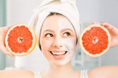 Hafif arka planda beyaz yüz maskesi ve greyfurt halfs ile güzel genç kadın. Elinde bir greyfurt tutan kafasına beyaz bir havlu ile mutlu kadın