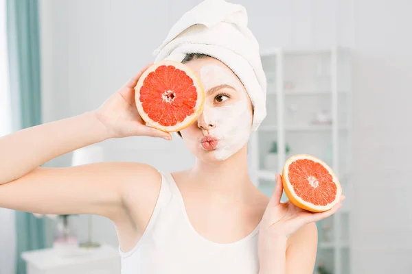 Portret van glimlachend mooi meisje met witte handdoek op het hoofdbedrijf helften van grapefruits in de buurt van het gezicht sluiten van een oog, een gezonde perfecte gladde huid, wit masker op de helft van het gezicht, over lichte achtergrond — Stockfoto