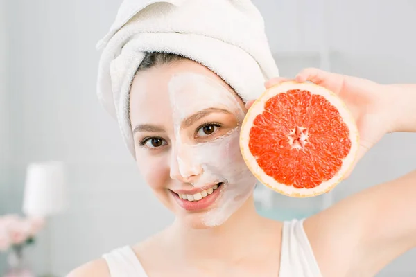 Retrato de jovem mulher bonita com brilho saudável pele lisa perfeita detém pedaço de toranja, toalha branca na cabeça, máscara branca no rosto. Cosméticos naturais, cuidados com a pele, conceito de tratamento facial . — Fotografia de Stock