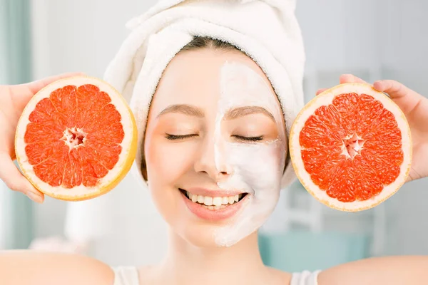 Gyönyörű fiatal nő, fehér arc maszk és a grapefruit halfs a világos háttér. Boldog asszony egy fehér törülközővel a fején kezében grapefruit a kezében — Stock Fotó