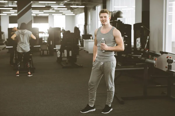 Rusttijd. Glimlachende jonge man in sportkleding staande met een fles water en kijken naar de camera op de achtergrond van de moderne fitness gym. — Stockfoto