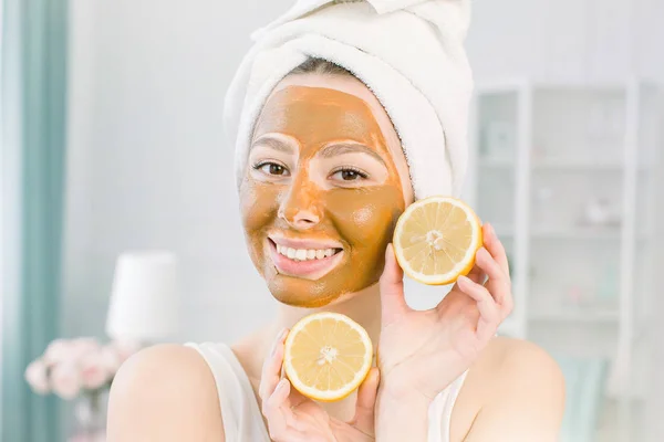 Beauty Skin Care Concept. Wanita Kaukasia yang menarik dengan handuk putih dan masker wajah lumpur bersenang-senang dengan dua bagian lemon, tembakan dalam ruangan di latar belakang cahaya — Stok Foto