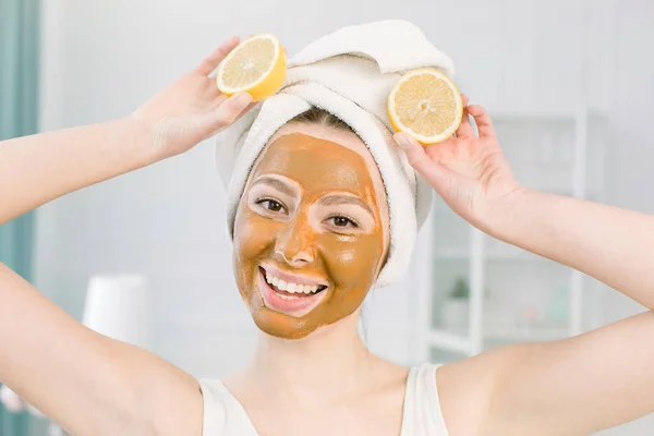 Beauty Skin Care Concept. Wanita Kaukasia yang menarik dengan handuk putih dan masker wajah lumpur bersenang-senang dengan dua bagian lemon, tembakan dalam ruangan di latar belakang cahaya — Stok Foto