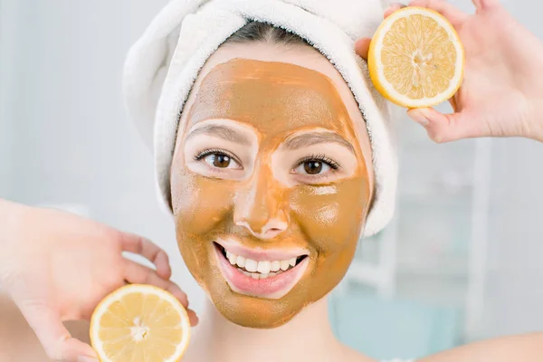 Beautiful young woman with facial mud mask and halfs of lemon on light background in spa center — Stock Photo, Image